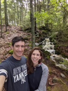 Two smiling adults stand in the woods next to a waterfall